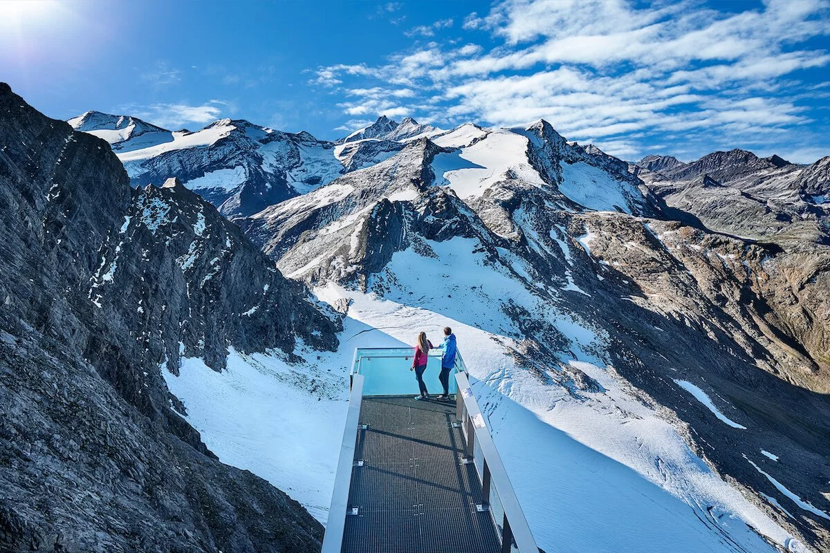 Aussichtsplattform in den österreichischen Alpen mit zwei Personen, umgeben von schneebedeckten Gipfeln und beeindruckender Hochgebirgslandschaft bei strahlendem Sonnenschein.