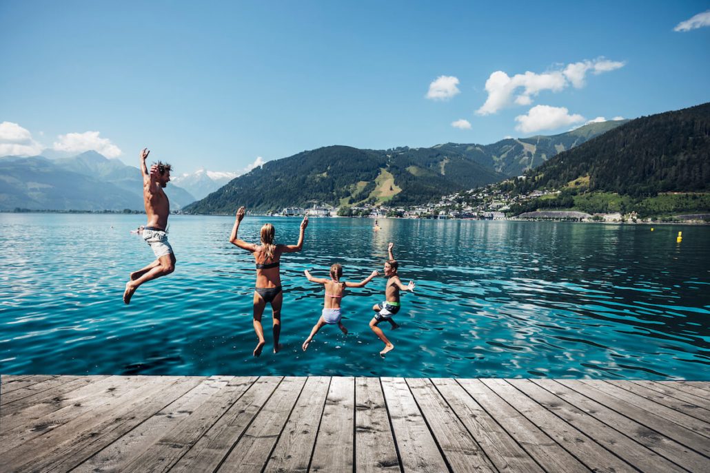 Familie springt von einem Steg in einen klaren Bergsee vor alpiner Kulisse