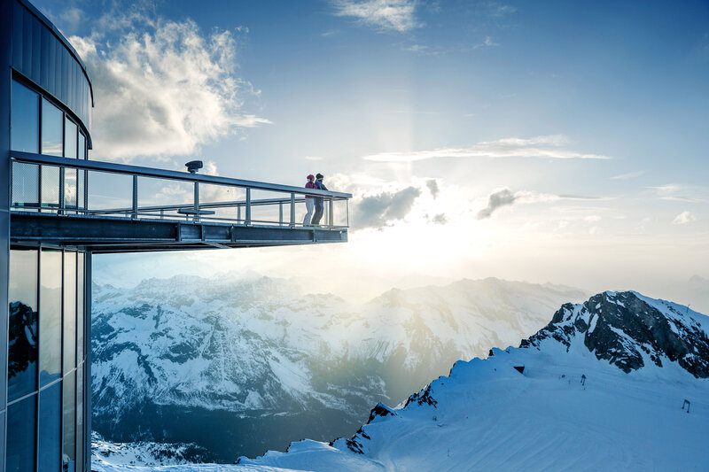 Zwei Personen auf einer verglasten Aussichtsplattform in schwindelerregender Höhe mit Blick auf verschneite Alpenberge.